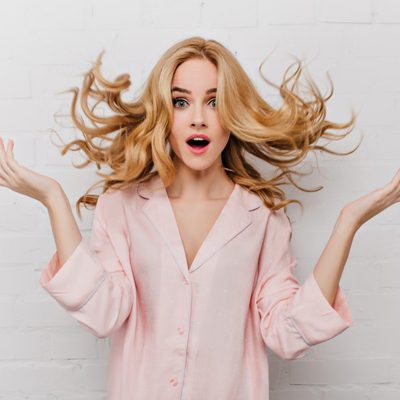 Ecstatic blue-eyed woman with long blonde hair posing in front of white bricked wall. Indoor shot of surprised girl in beautiful pink pyjamas.